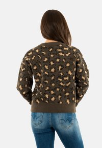Donna con lunghi capelli castani che indossa un maglione leopardato marrone e beige e jeans blu, voltata di spalle davanti a uno sfondo neutro.