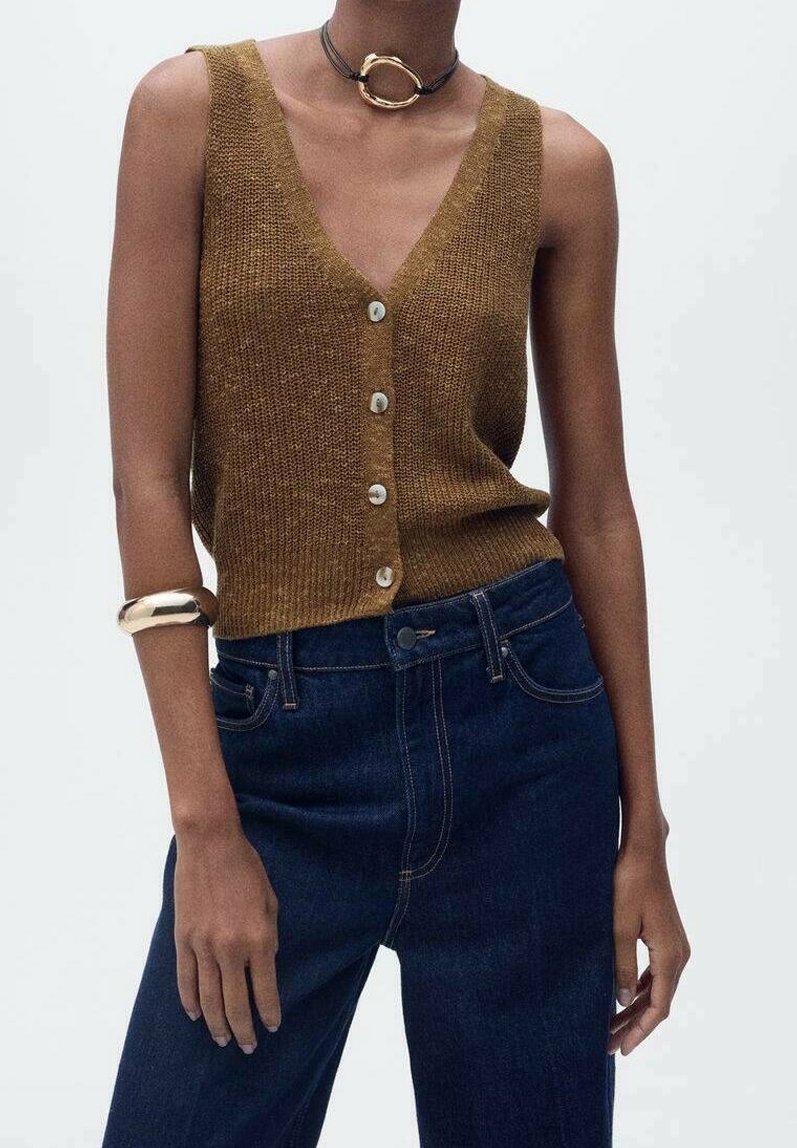 Brązowy, dziany top bez rękawów z głębokim dekoltem w kształcie V i zapinaniem na guziki, w połączeniu z ciemnoniebieskimi jeansami o wysokim stanie i masywną bransoletką.