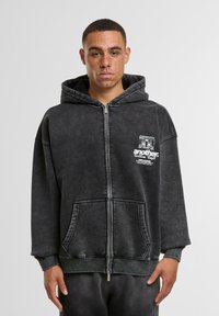 Grauer Reißverschluss-Hoodie aus weichem Material, mit einem Logo auf der Vorderseite, großer Tasche und gerippten Bündchen, mit lässigem Schnitt und übergroßem Design.