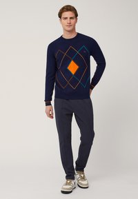 Maglione blu navy con motivo geometrico in arancione, marrone e teal; polsini a coste con strisce a contrasto; abbinato a pantaloni scuri e sneakers.