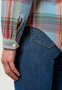 Multifärgad rutig skjorta med röda, gröna och blå ränder; knappmanschettdetalj synlig. Bärs med mörkblå denimjeans, där fickan syns.