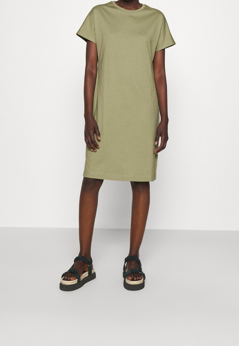 Personne portant une robe courte à manches courtes de couleur vert olive et des sandales compensées noires à larges brides, debout devant un fond uni.