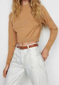 Gekürzter, beige Pullover mit hohem Kragen und eng anliegenden Bündchen, kombiniert mit hoch taillierten, hellen Jeans und einem braunen Gürtel. Glatte Textur.