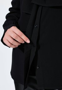 Sort bluse med knaplukning, blød tekstur og lange ærmer. Detalje af en hånd, der peger på et knapområde med et plisseret frontdesign.