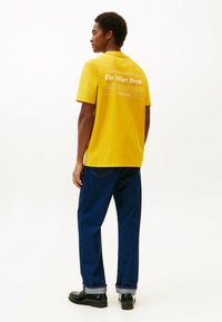 Junger Mann, der seitlich steht, trägt ein gelbes T-shirt mit weißem Text auf dem Rücken, dunkle Jeans und schwarze Schuhe vor einem schlichten weißen Hintergrund.