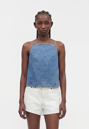 Mujer con el cabello trenzado que lleva una camiseta sin mangas de mezclilla azul y pantalones cortos blancos con cordón, mirando hacia adelante sobre un fondo liso.