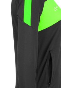 Giacca nera con accenti in rete verde brillante, tasca con zip, vestibilità slim e texture liscia, evidenziando un design a colori contrastanti.