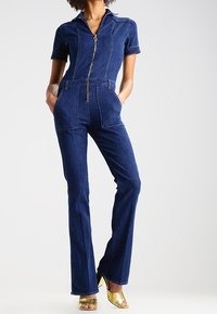 Jeansbyxa i mörkblått med korta ärmar, dragkedja fram och sidofickor. Har en åtsittande bodice och design med utsvängda ben.