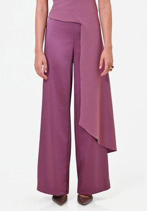 Femme portant un pantalon violet à jambes larges avec un panneau en tissu drapé asymétrique et des chaussures brunes pointues, debout sur fond blanc.