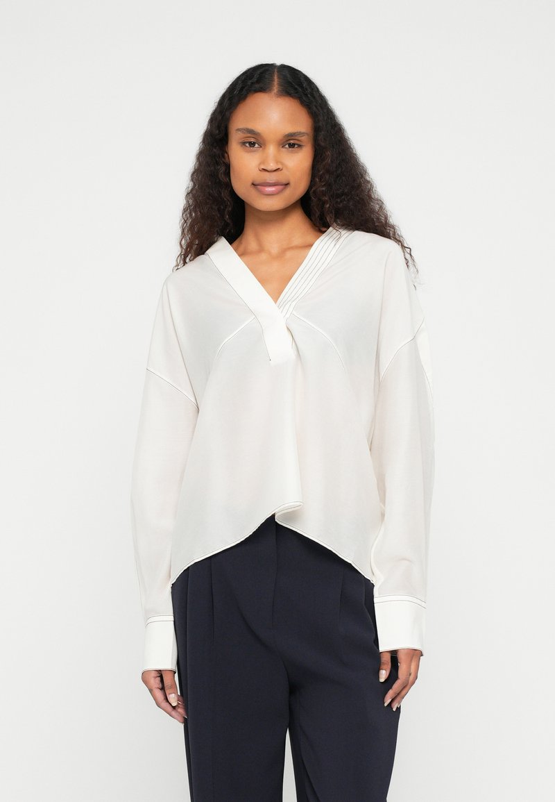 Hvit, lett bluse med v-hals og plissert detalj ved halsen. Har oversized ermer og buet kant, stylet casual.