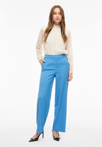 Světle krémová blůza s texturovanými tečkami a volánkovým výstřihem, v kombinaci s zářivě modrými širokými kalhotami a černými lodičkami na vysokém podpatku se zlatými akcenty.