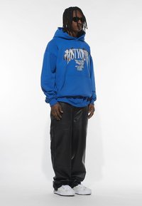 Felpa blu con cappuccio e stampa grafica, abbinata a pantaloni di pelle neri e sneakers bianche. Modello con occhiali da sole.