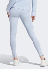 Persona vestida con leggings azul claro con rayas blancas a los lados y zapatillas blancas, de pie con las piernas separadas frente a un fondo liso.