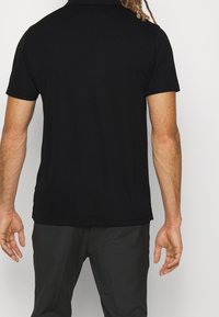 Mężczyzna z dredami, ubrany w prostą czarną koszulę z krótkim rękawem i ciemne spodnie, widziany z tyłu na jednolitym tle.