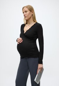 Έγκυος γυναίκα με μαύρο μπλουζάκι μακριάς μανίκας και γκρι παντελόνι κρατά ασημένιο τσαντάκι με το ένα χέρι, ενώ το άλλο ακουμπά στην κοιλιά της, στέκεται μπροστά σε λευκό φόντο.