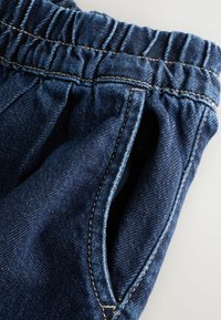 Pantalones de mezclilla de color azul oscuro, con cintura elástica y dos bolsillos laterales. Presentan costuras visibles en un color claro contrastante.