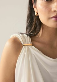 White draped fabric top with a gold hardware accent on the shoulder, showcasing soft texture and elegant folds. Close-up view of the shoulder area.