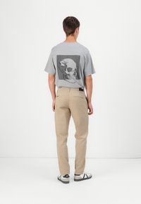 Šedé tričko s krátkým rukávem, na kterém se na zádech nachází velká černá grafika lebky. Spárované s khaki kalhotami a černobílými teniskami.