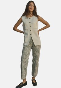 Gilet beige senza maniche con due tasche frontali e chiusura a bottoni, abbinato a pantaloni beige con stampa serpente e ballerine nere.