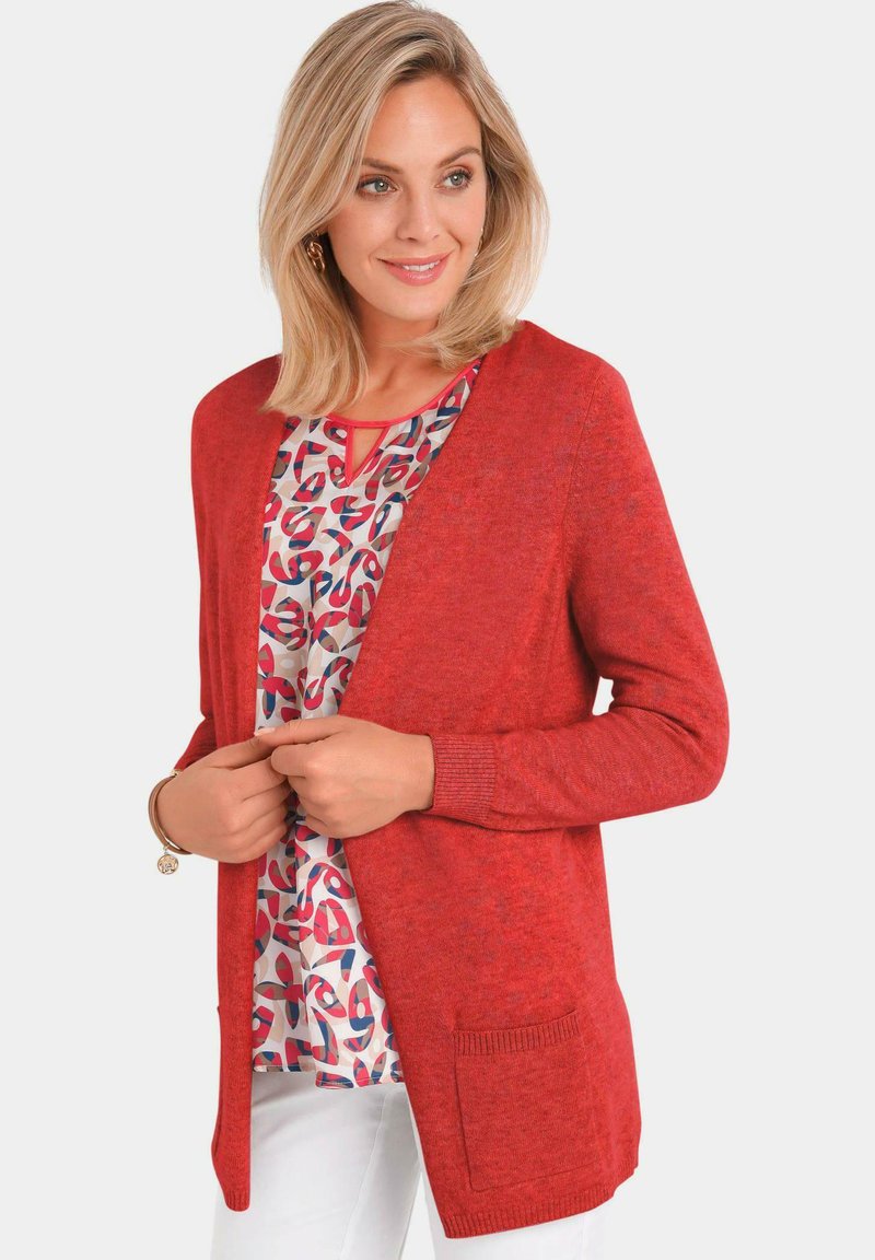 Cardigan rouge en tissu tricoté, avec une coupe décontractée, un front ouvert, des manches longues et une poche avant. Superposé à un chemisier à motifs.