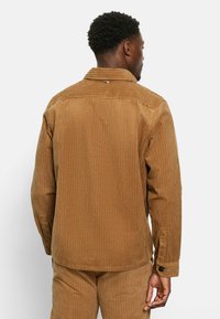 Braune Cordjacke mit vertikalen Rippungen, ausgestattet mit einem umklappbaren Kragen, Knopfmanschetten und einem abgerundeten Saum. Die Rückansicht zeigt die Texturdetails.