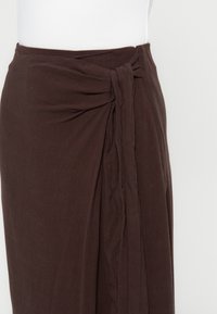 Jupe portefeuille marron foncé avec détail de tissu froncé et nœud noué à la taille sur un mannequin blanc.
