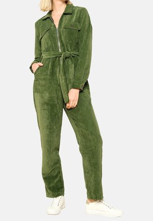 Kobieta w zielonym kombinezonie sztruksowym z przednim zamkiem błyskawicznym, wiązanym paskiem w talii, kieszeniami i białymi trampkami na gładkim tle.