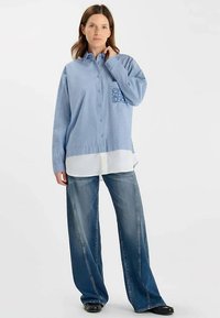 Chemise bleue claire boutonnée avec un motif texturé et une poche latérale, portée sur une chemise blanche, associée à un jean bleu délavé à jambes larges.