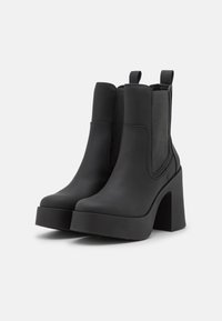 Botines negros hasta el tobillo con un tacón grueso, hechos de cuero suave y paneles laterales elásticos para facilitar su uso. Presentan una suela de plataforma y un diseño de punta redonda.