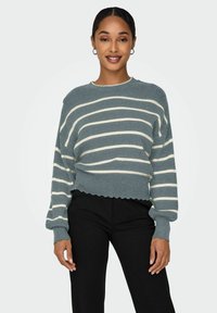 Pull en maille rayé avec une texture côtelée en bleu sarcelle et des rayures crème, avec des manches larges et un ourlet festonné, associé à un pantalon noir.