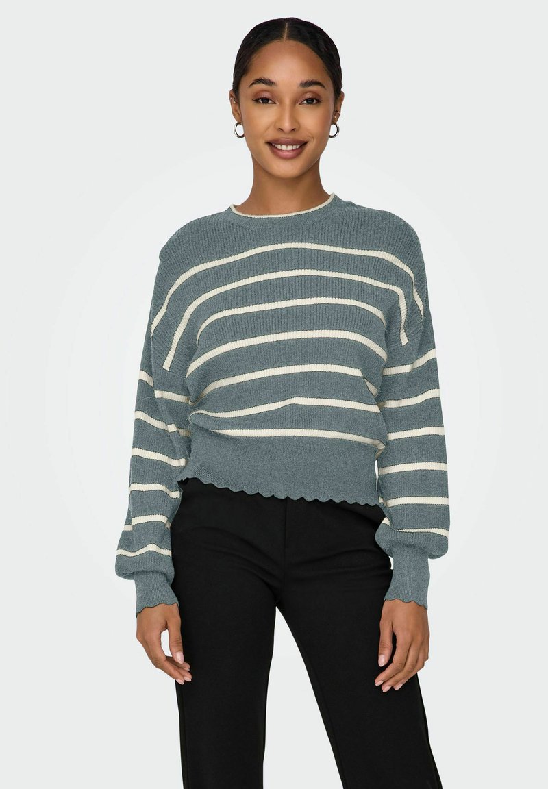 Pull en maille rayé avec une texture côtelée en bleu sarcelle et des rayures crème, avec des manches larges et un ourlet festonné, associé à un pantalon noir.