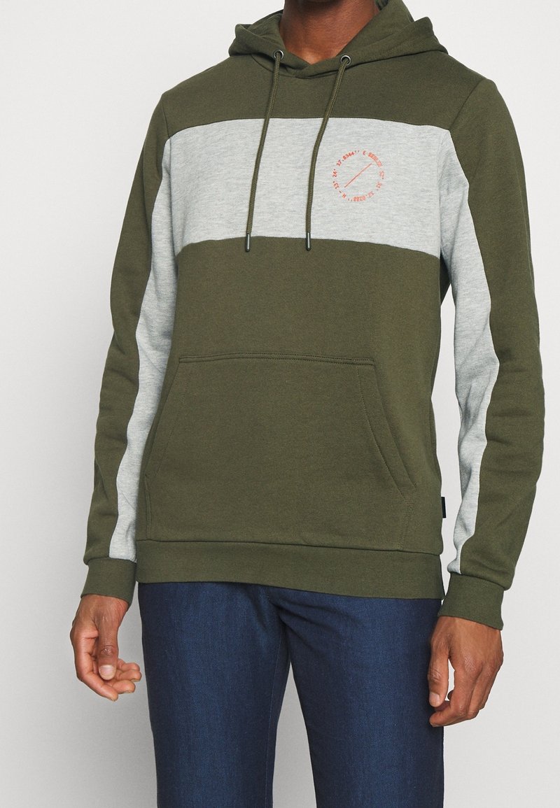 Homme portant un sweat à capuche color block vert olive et gris clair avec poche avant et petit logo rouge circulaire sur la poitrine.