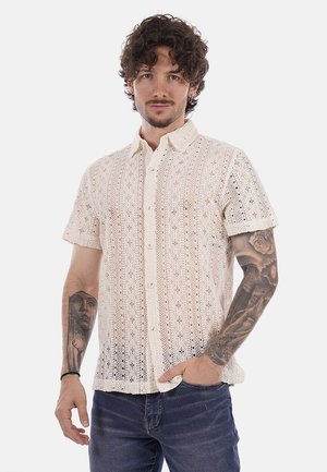 Uomo con capelli ricci che indossa una camicia bianca in pizzo a maniche corte con bottoni e jeans blu, braccia tatuate visibili, in piedi contro uno sfondo semplice.
