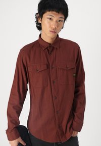 Camisa de botão de manga longa na cor rust com dois bolsos no peito, botões pretos e uma bainha arredondada. O tecido tem uma textura suave.