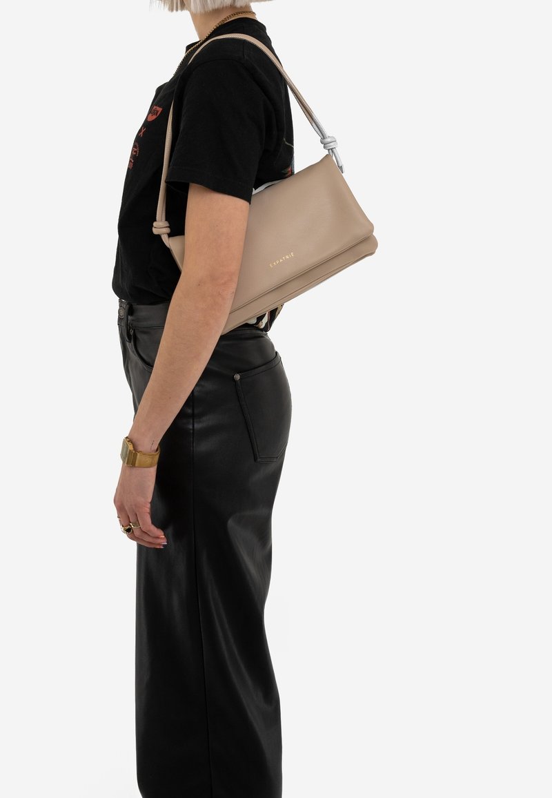 Femme portant un t-shirt noir et une jupe en cuir, portant un sac à bandoulière beige avec un texte doré sur un fond uni.