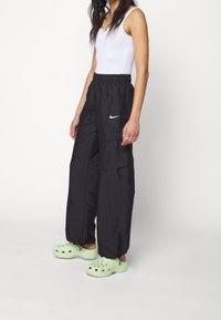 Zwarte cargobroek met elastische taille, voorzien van een wit Nike-logo, gecombineerd met groene klompen. De broek heeft een losse pasvorm en taps toelopende boorden.