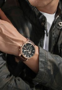 Uomo che indossa un orologio cronografo con cinturino in pelle marrone e quadrante nero, vestito con una giacca mimetica e una camicia bianca.