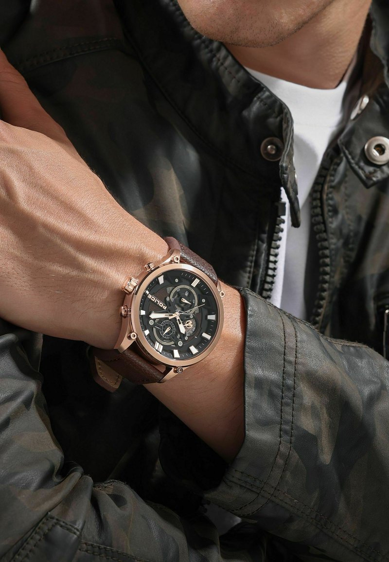 Uomo che indossa un orologio cronografo con cinturino in pelle marrone e quadrante nero, vestito con una giacca mimetica e una camicia bianca.