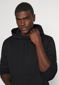 Svart hoodie med dragsko, mjukt tyg, framficka och ribbade ärmslut. Modellen poserar med handen på kragen för att visa strukturen och passformen.