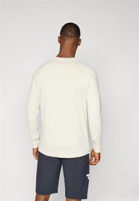 Vaaleanbeige pitkähihaisessa paidassa on raglan-hihat, ja se on yhdistetty tummansinisiin shortseihin, joissa on valkoinen logo vasemmassa lahkeessa. Pintakuvio on sileä.