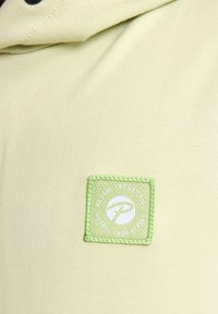 Tessuto giallo chiaro con una texture liscia presenta un'etichetta ricamata verde con un logo, inserita in una patch rettangolare sul petto.