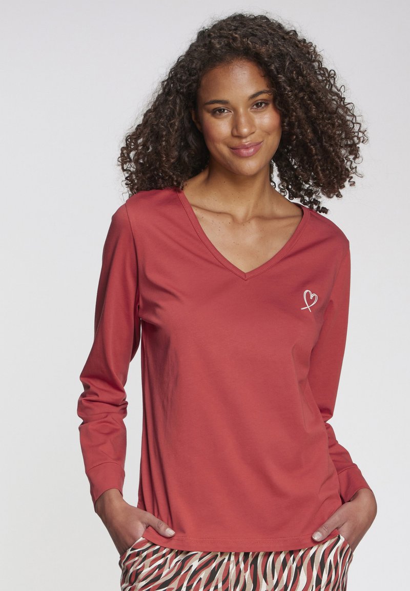 Rotes Langarmshirt mit V-Ausschnitt, versehen mit einer weißen Herzstickerei auf der linken Brust, gecombineerd mit gemusterten Hosen. Weiche Stofftextur.