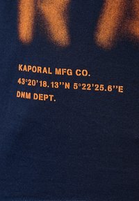 T-shirt navy avec un graphisme orange : "KAPORAL MFG CO." et des coordonnées accompagnées de "DNM DEPT." imprimé dans une police audacieuse et moderne.