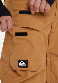 Braune Schneehosen aus einem glatten, strapazierfähigen Stoff. Verfügt über Klettverschluss-Taschen und ein schwarzes Logo-Patch. Eingenähte Details erhöhen die Funktionalität.