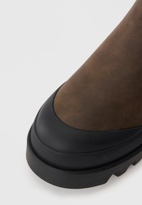 Bottine en cuir marron avec une semelle en caoutchouc noire, présentant une finition texturée et un bout arrondi, alliant durabilité et design moderne.