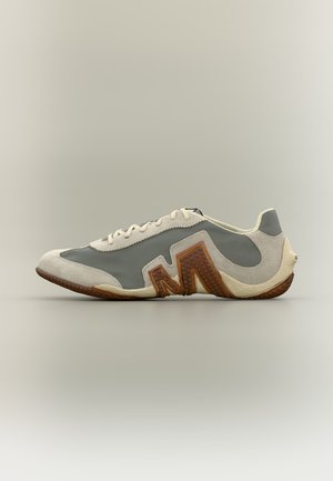 Sneaker χαμηλού προφίλ με μπεζ καστόρι και γκρι ύφασμα, με μεγάλο καφέ υφή "M" στο πλάι και λευκά κορδόνια.
