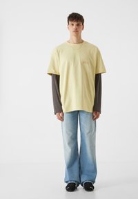 Geltona per didelė marškinė su pilku raukšlėtu ilgomis rankovėmis, derinami su šviesiai mėlynomis plačiomis džinsinėmis kelnėmis ir juodais sportbačiais.