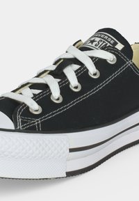 Baskets basses Converse noires avec lacets blancs, semelle en caoutchouc blanche et logo visible sur la languette, sur fond uni.