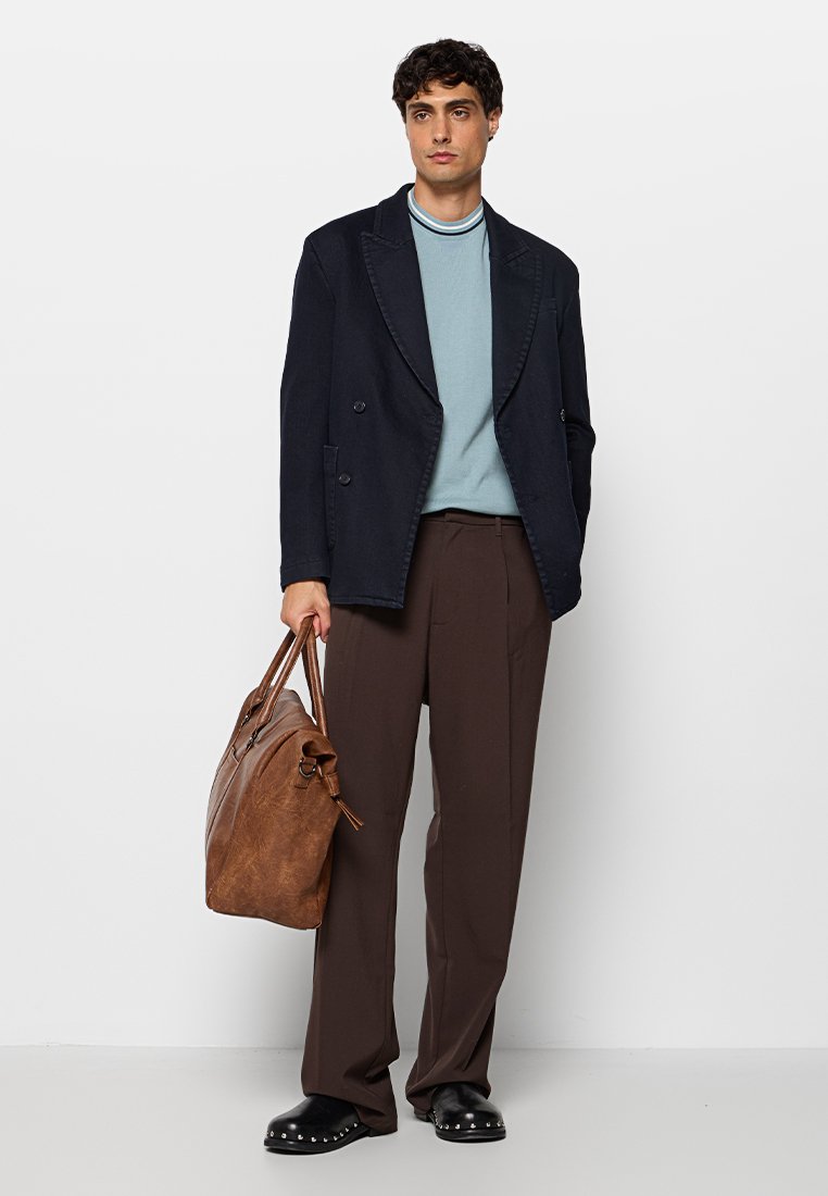 Blazer blu navy doppiopetto sopra un maglione azzurro chiaro, pantaloni larghi marroni e scarpe nere slip-on, con una borsa in pelle marrone.