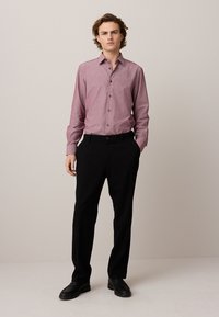 Giovane uomo in piedi con le mani in tasca che indossa una camicia bordeaux con bottoni, pantaloni neri e scarpe nere su uno sfondo semplice.